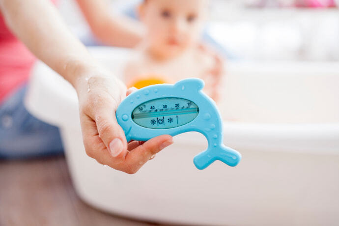 Bebê tomando banho e mulher segurando termômetro de banheira em formato de golfinho.