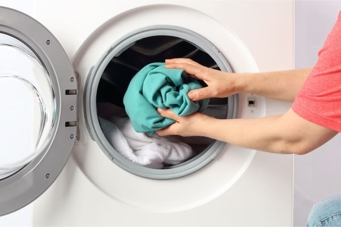 Indivíduo colocando uma peça de roupa verde dentro da máquina de lavar.