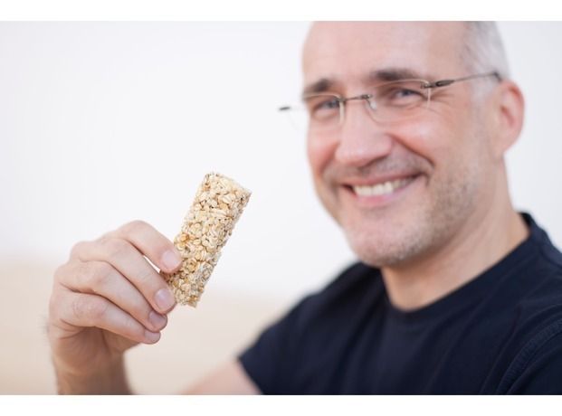 Homem de meia idade, com óculos de grau, sorrindo e segurando uma barra de cereal 