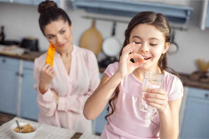 Criança tomando vitamina com a mãe feliz ao fundo.
