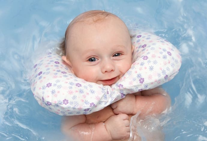 Bebê sorrindo na água com almofada para banho em volta do pescoço.