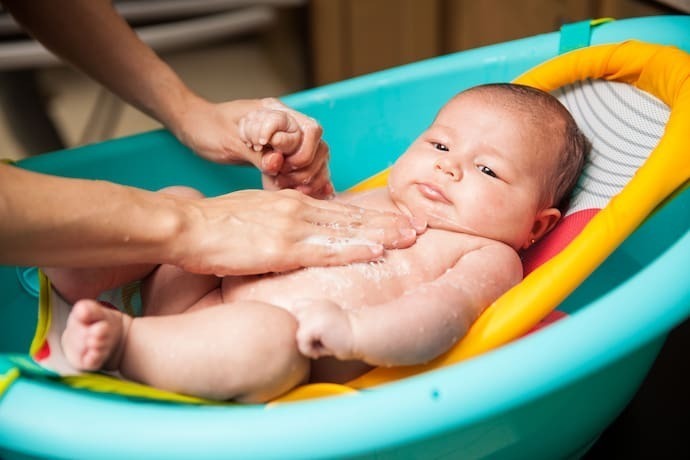 Bebê em banheira verde-água com almofada para banho laranja.