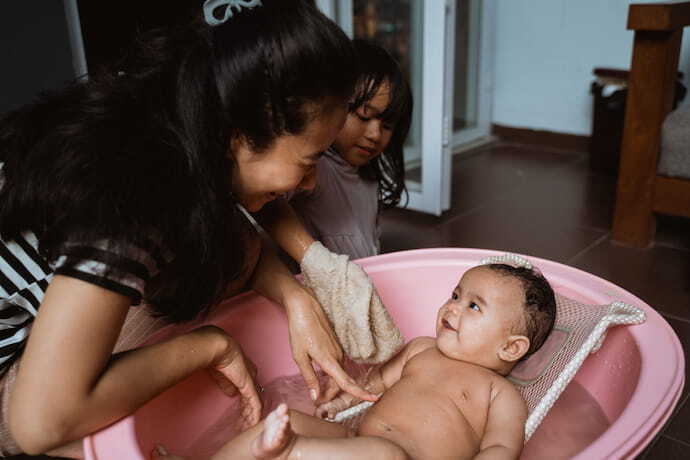 Bebê sorrindo para mãe em banheira rosa com almofada para banho branca.