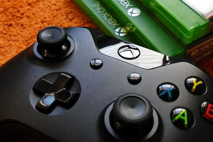 Controle de Xbox One preto com jogos ao fundo.