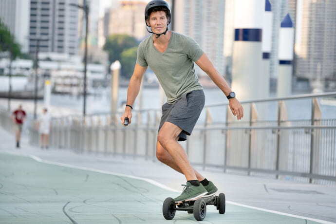 Homem de capacete andando em skate elétrico segurando controle remoto.