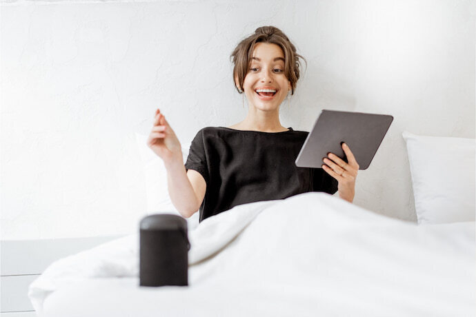 Mulher sorrindo com smart speaker na cama e tablet na mão.