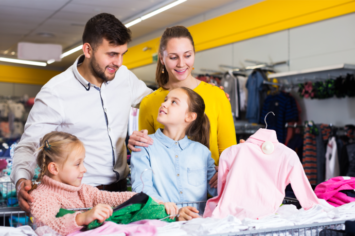 Pai, mãe e duas filhas escolhendo roupa na loja. Uma das filhas segurando camisa rosa na mão.
