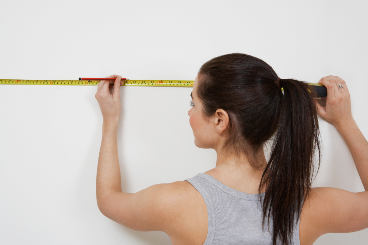 Mulher medindo a parede com uma fita métrica e um lápis.