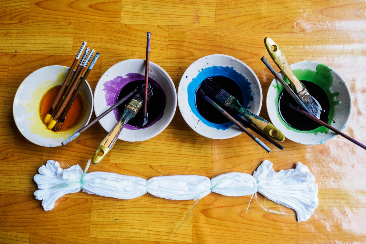 Quatro recipientes com tinta, pincéis e tecido branco sobre a mesa de madeira.