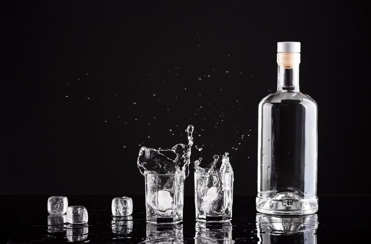 Uma garrafa de vidro transparente com dois copos com vodka e gelo ao lado em um fundo preto