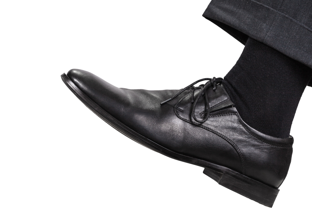 Perna esquerda masculina em sapato preto de bico fino, em um fundo branco.