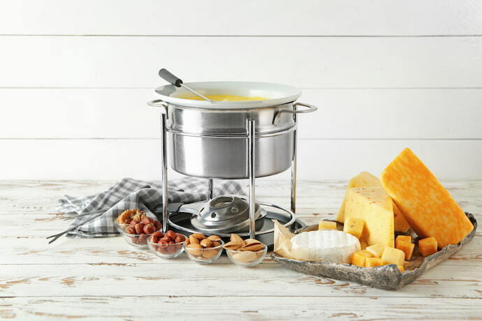 Aparelho de fondue prata com fogareiro e queijo ao redor.