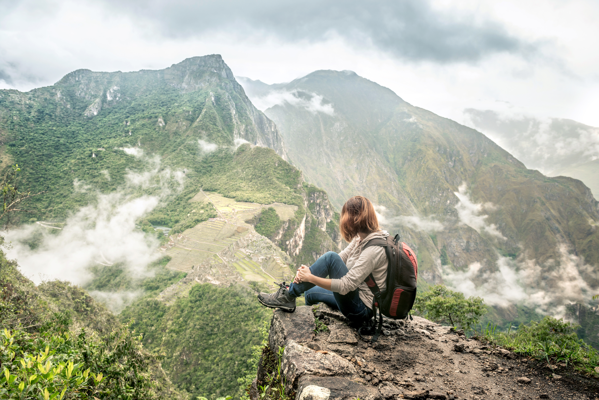 Mulher com mochila, botas e roupas confortáveis sentada próxima a um abismo de onde se vê várias montanhas peruanas, como a Montanha Machu Picchu