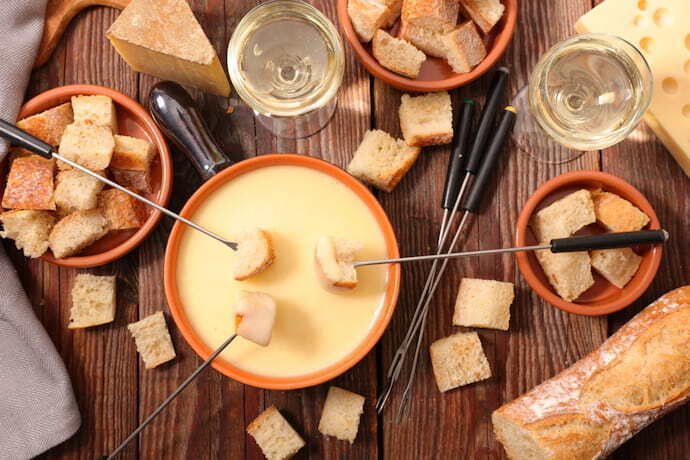 Fondue de cerâmica cheia de queijo e pães, vinhos e queijos ao redor.