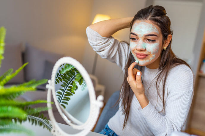 Mulher aplicando creme esfoliante verde no rosto, em frente a um espelho.