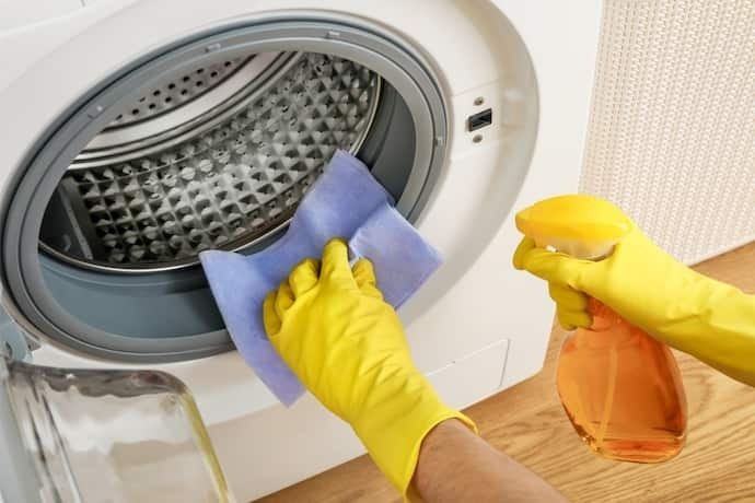 Indivíduo com luvas e produto de limpeza, higienizando o interior da máquina de lavar.