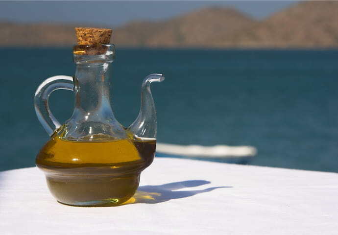 Azeite de oliva em pequena jarra sobre mesa, ao fundo se vê o mar