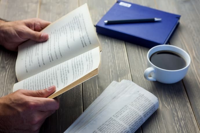 Pessoa lendo livro apoiado na mesa com copo de café 