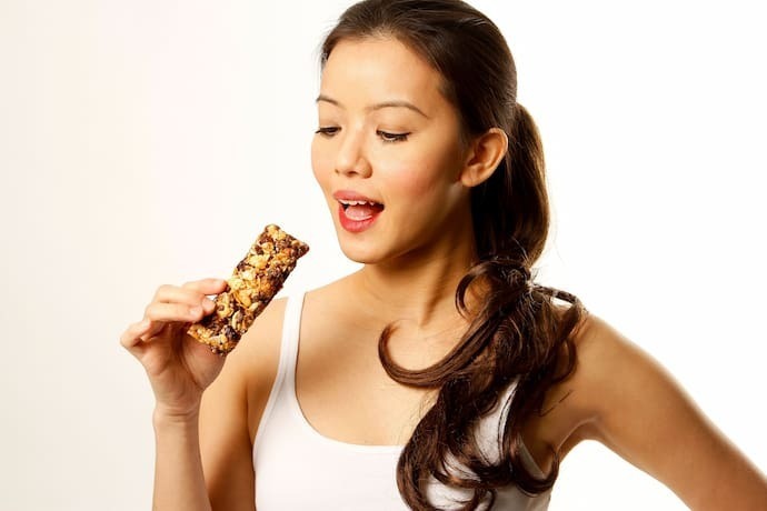 A imagem apresenta uma mulher jovem com a boca semi aberta e segurando uma barra de cereal.