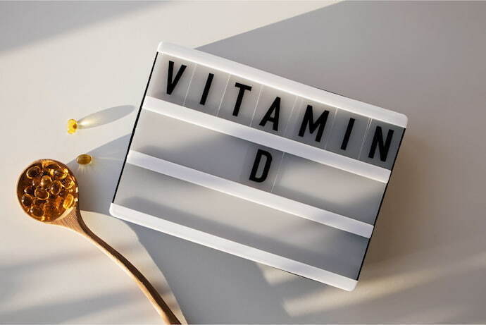 Placa escrito "Vitamina D" em inglês com cápsulas de vitamina ao redor em fundo branco.