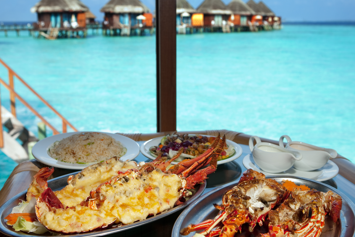 Mesa com pratos como lagosta, molhos, arroz, etc; ao fundo se vê o mar cristalino e alguns bangalôs